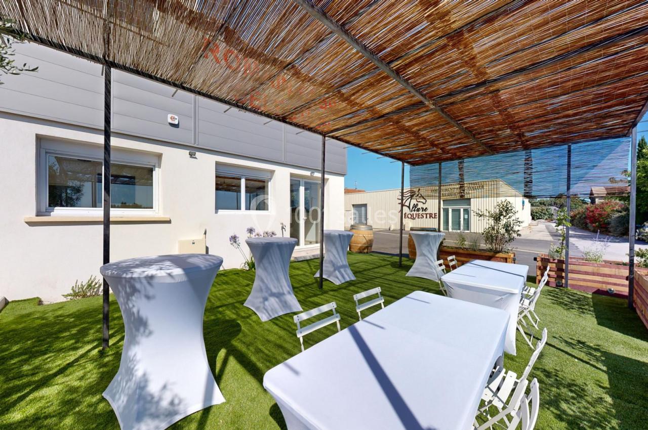 Terrasse extérieure avec tables et chaises blanches sous une pergola en bois, entourée de verdure et de bâtiments.