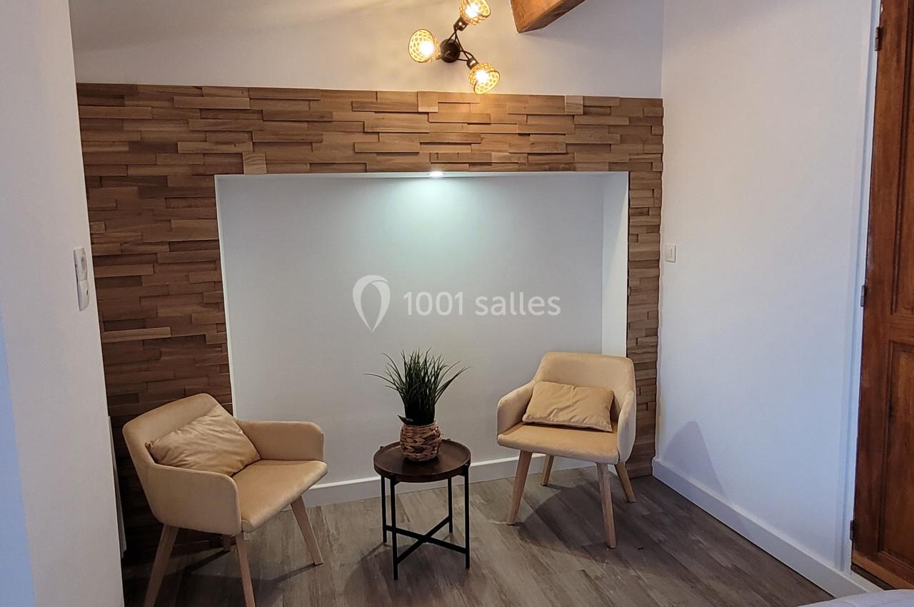 Deux fauteuils beiges entourent une petite table avec une plante, dans un coin décoré de bois et éclairé par une applique…