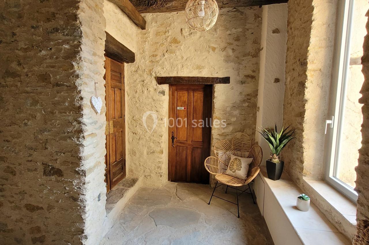 Couloir en pierre avec plafond en bois, porte en bois, fauteuil en rotin, plante et lumière naturelle provenant d'une…