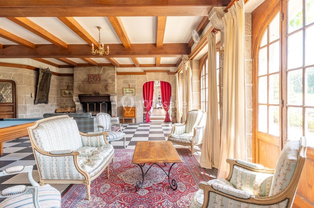 Salon lumineux avec poutres apparentes, cheminée en pierre, mobilier classique et grandes fenêtres donnant sur l'extérieur.