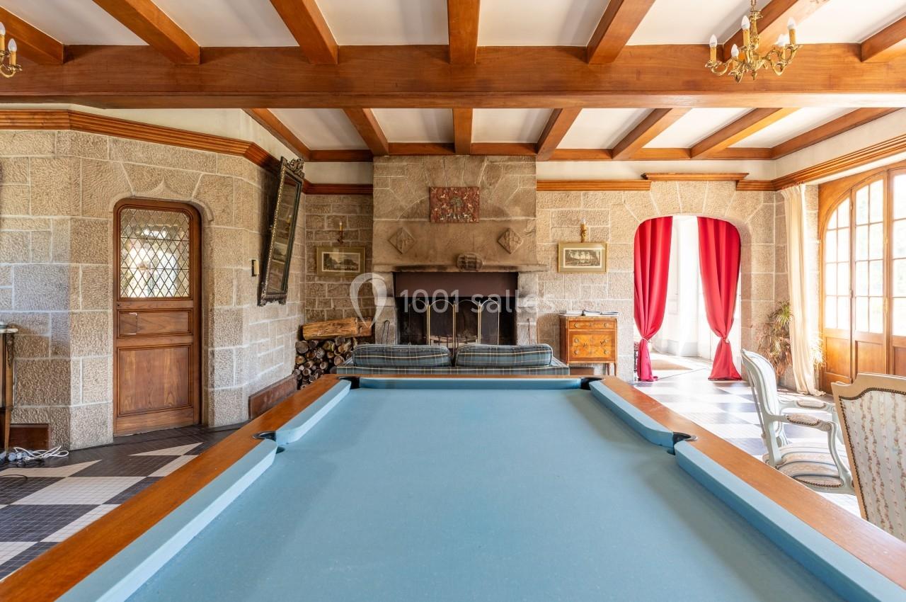 Salle avec table de billard, cheminée en pierre, poutres apparentes et grandes fenêtres avec rideaux rouges.