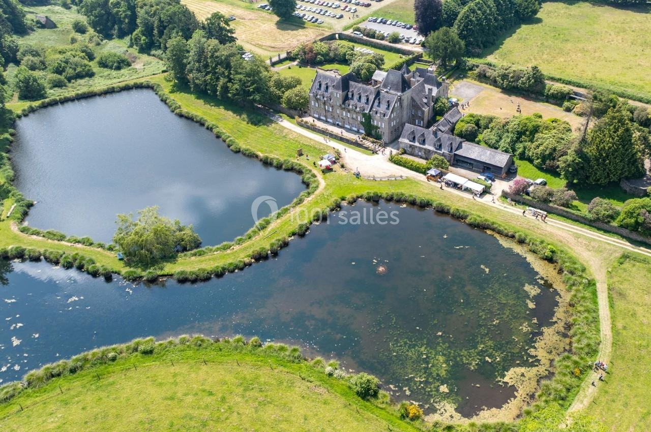 Vue aérienne d'un manoir entouré de deux étangs et de verdure, avec un parking visible en arrière-plan.