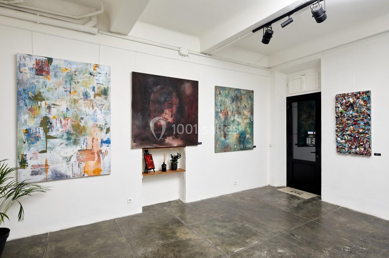 Galerie d'art avec plusieurs tableaux abstraits accrochés sur des murs blancs, éclairés par des spots au plafond.