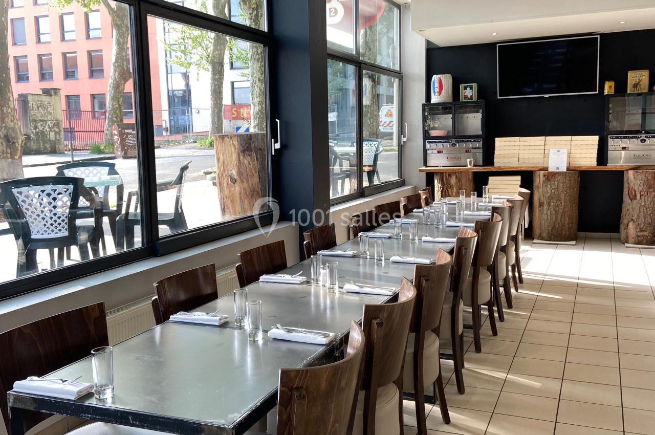 Salle de restaurant lumineuse avec tables alignées, chaises en bois et grandes fenêtres donnant sur l'extérieur.