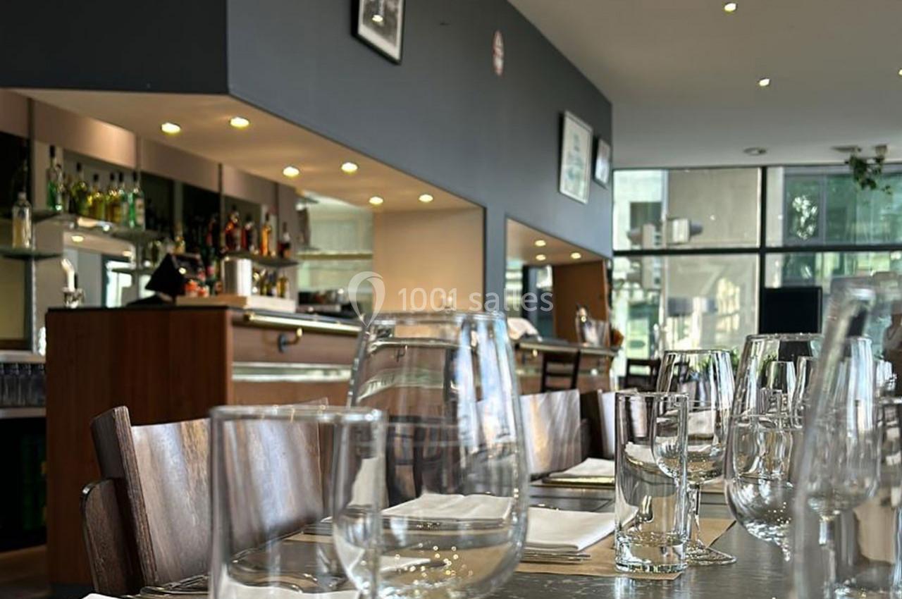 Salle de restaurant moderne avec tables dressées, verres et serviettes, vue sur un bar et de grandes baies vitrées.