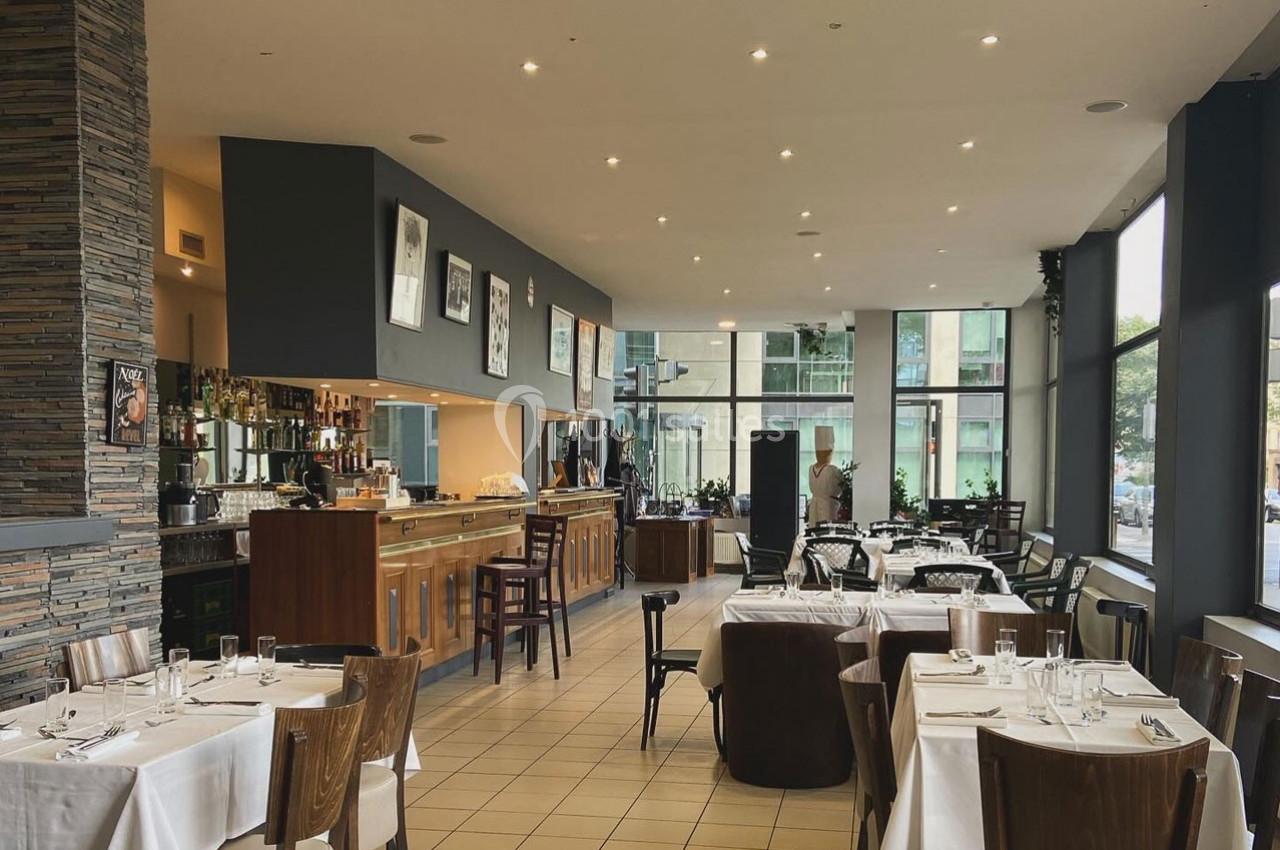 Salle de restaurant lumineuse avec tables dressées, bar en bois et grandes baies vitrées donnant sur l'extérieur.