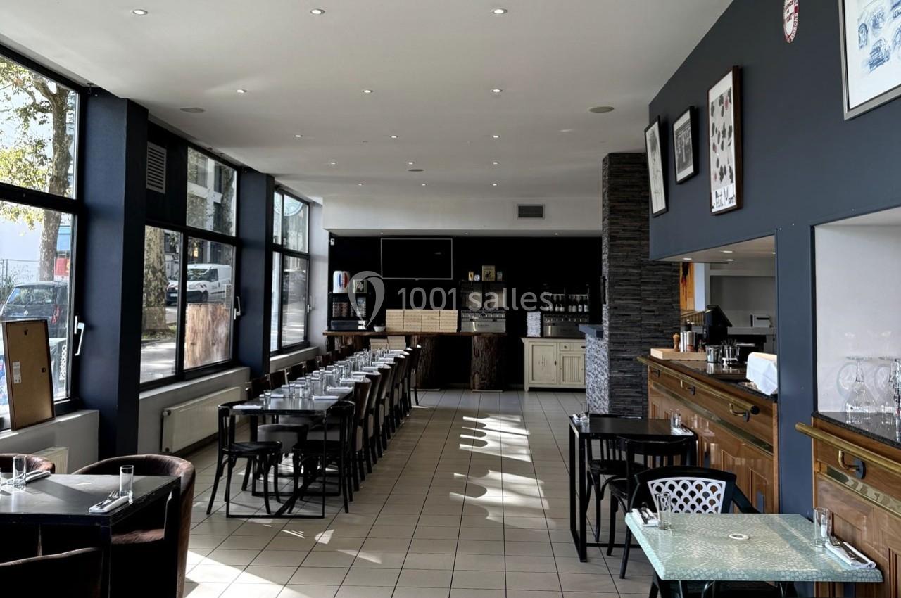 Salle de restaurant lumineuse avec tables dressées, grandes baies vitrées et comptoir en bois à droite.