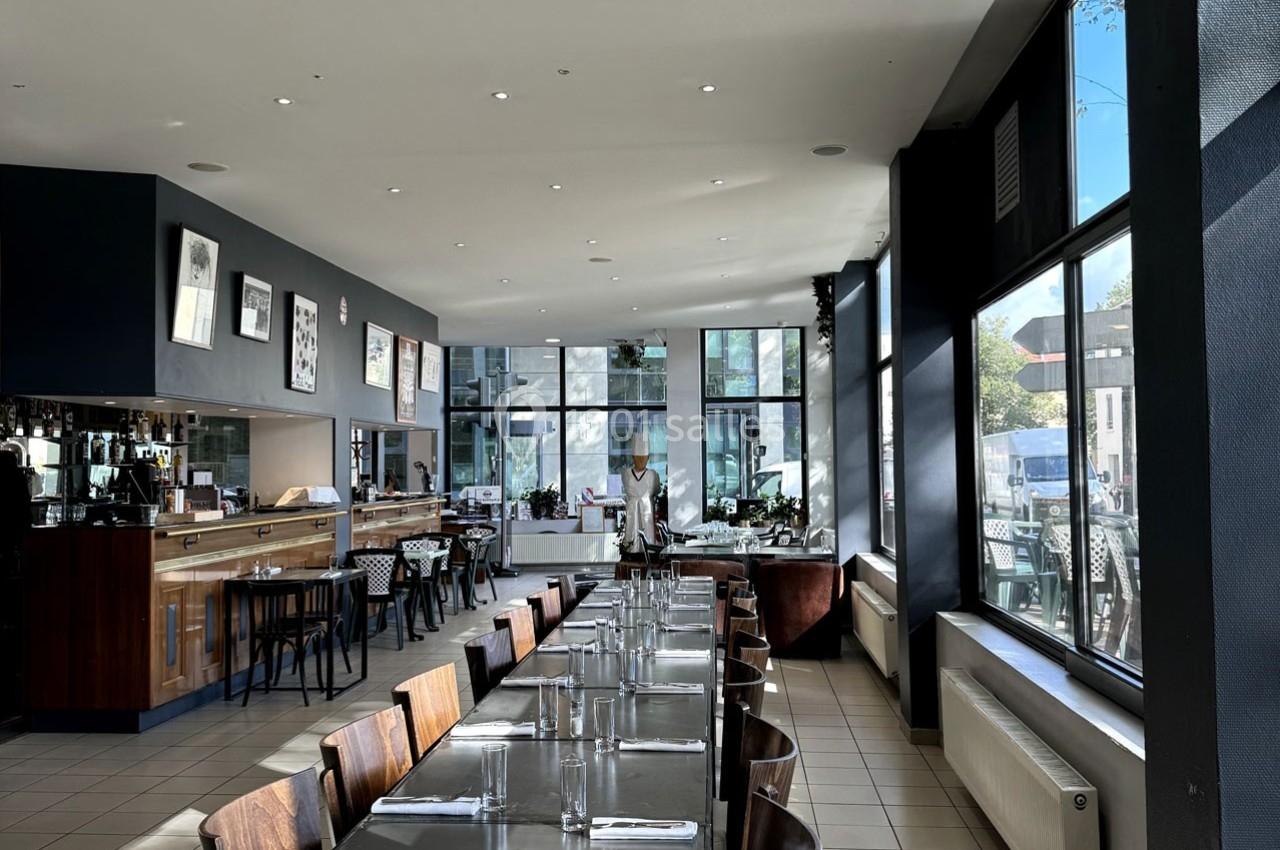 Salle de restaurant lumineuse avec tables dressées, grandes baies vitrées et un comptoir en bois au fond.
