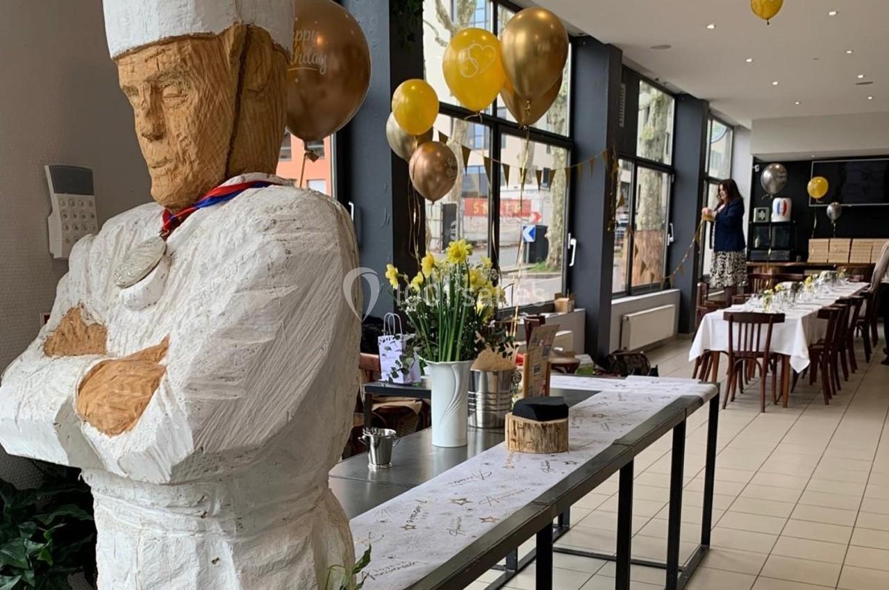 Statue en bois d'un chef cuisinier dans une salle décorée avec des ballons dorés et des tables dressées.