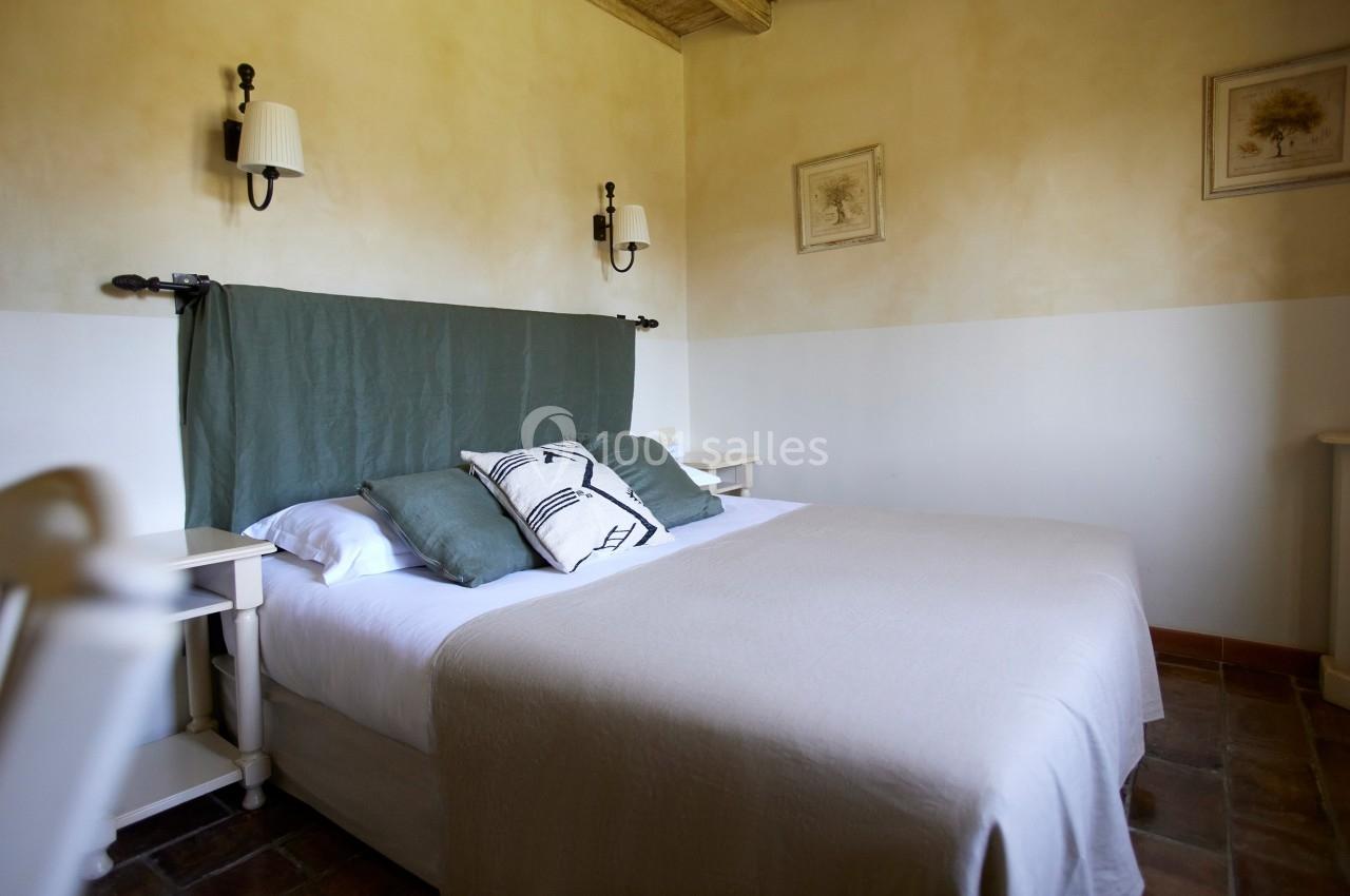 Lit double avec draps beiges, coussins verts et noirs, dans une chambre lumineuse aux murs clairs et décor minimaliste.