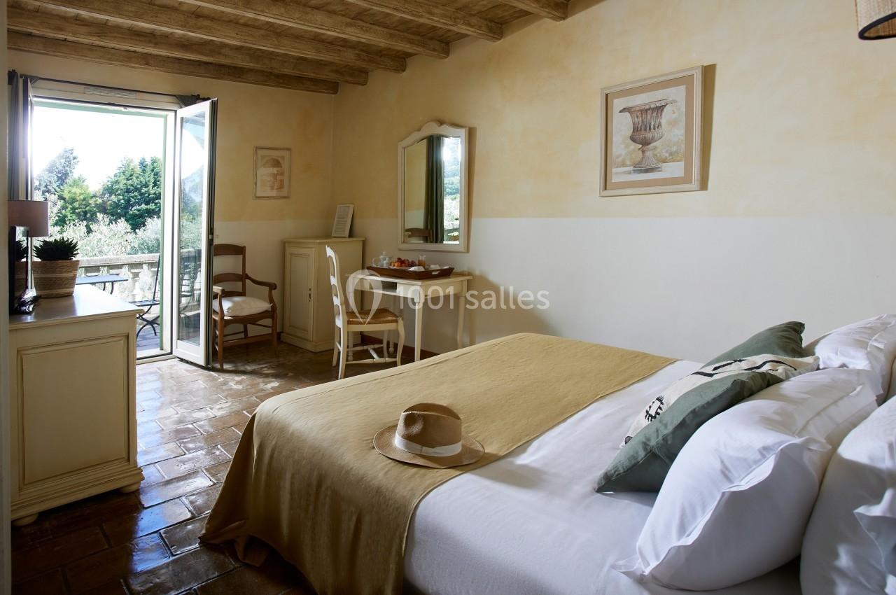 Chambre lumineuse avec lit double, bureau, chaise et accès à un balcon offrant une vue sur un jardin verdoyant.