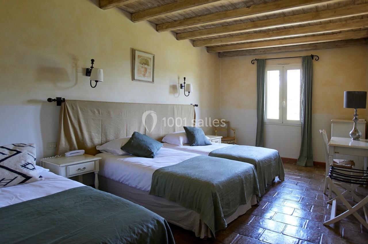 Chambre lumineuse avec trois lits simples, murs clairs, poutres apparentes, fenêtre avec rideaux verts et mobilier sobre.