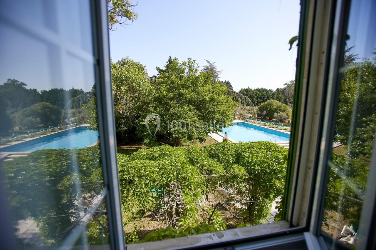 Vue depuis une fenêtre sur un jardin verdoyant avec deux piscines entourées de végétation sous un ciel dégagé.