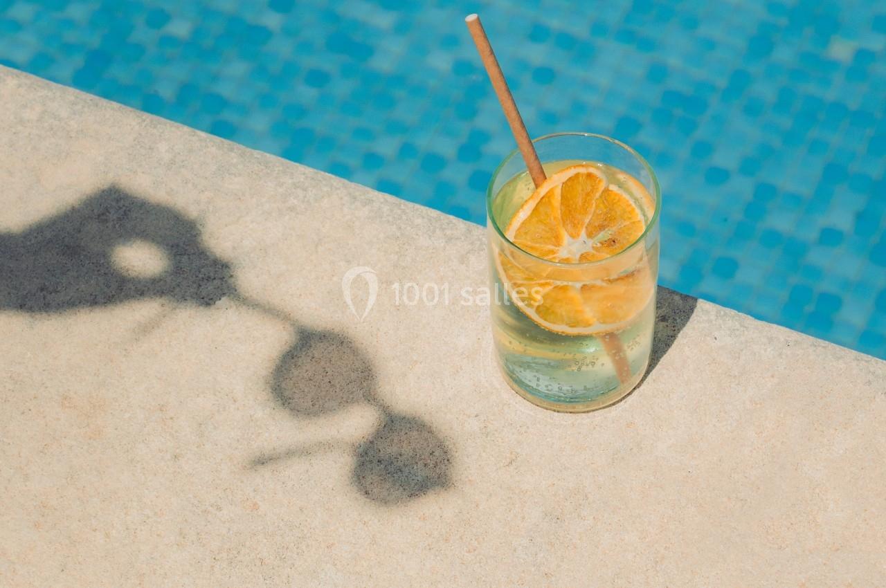 Verre de boisson fraîche avec des tranches d'orange et une paille, posé sur le bord d'une piscine en mosaïque bleue.