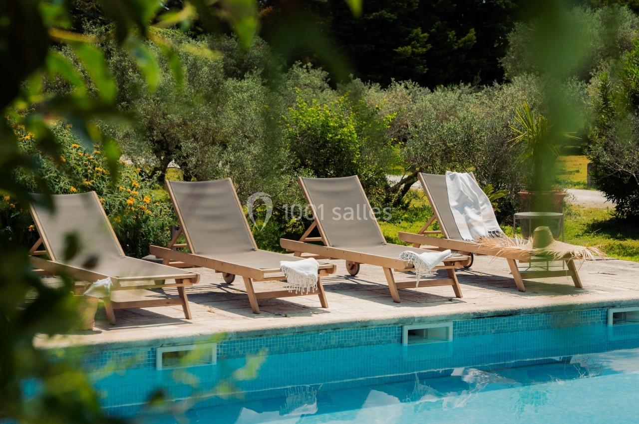 Chaises longues alignées au bord d'une piscine, entourées de végétation et d'arbres sous un ciel dégagé.