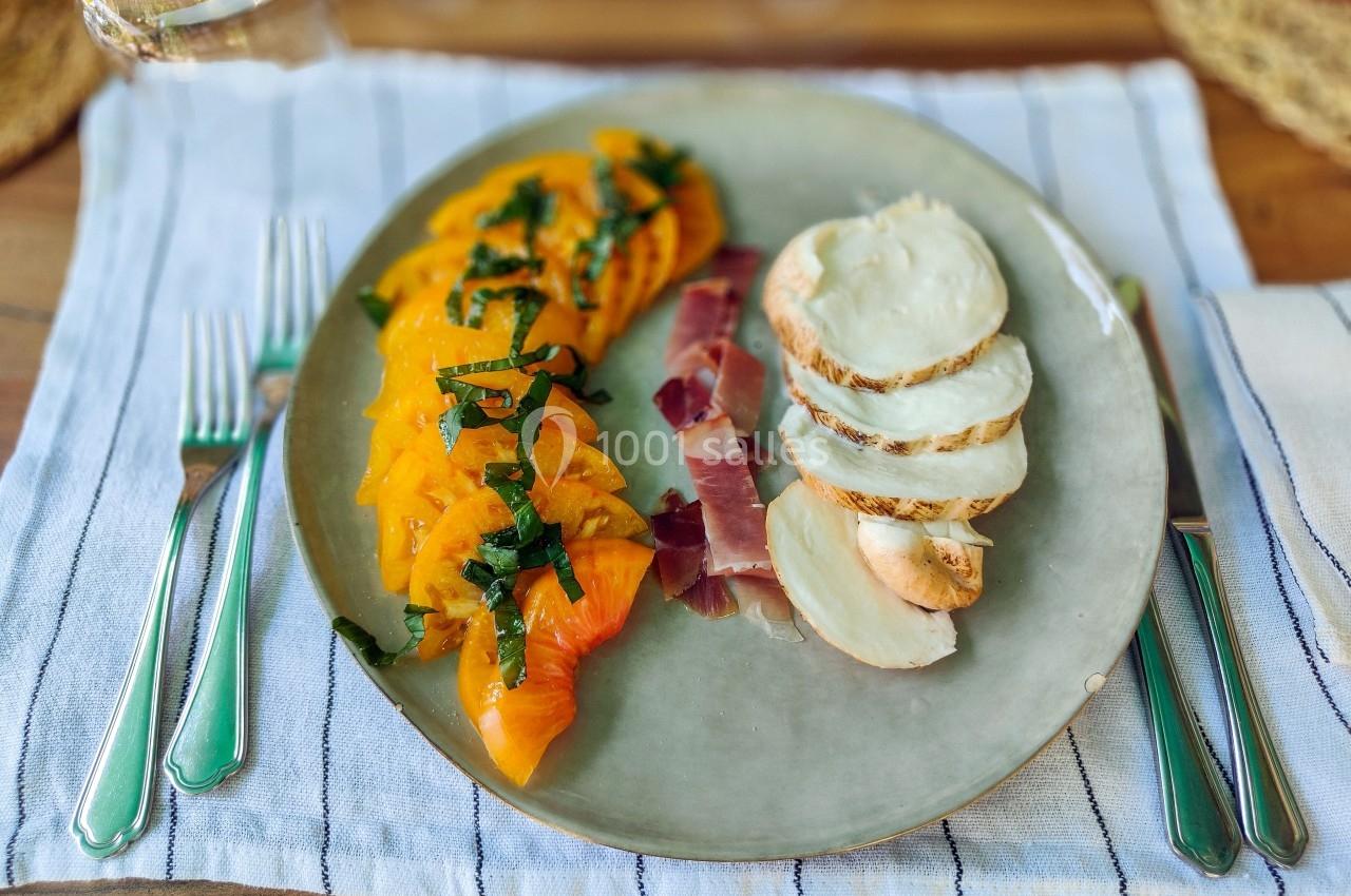 Assiette contenant des tranches de tomates jaunes, mozzarella, jambon cru et basilic, sur une nappe rayée.