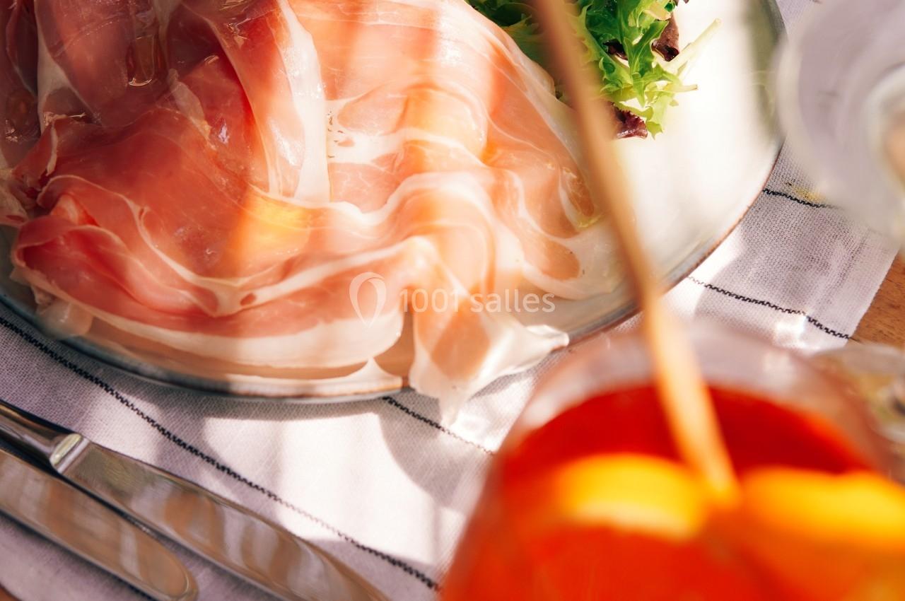 Assiette de jambon cru accompagnée de salade verte, servie sur une nappe rayée avec un verre de cocktail orange.