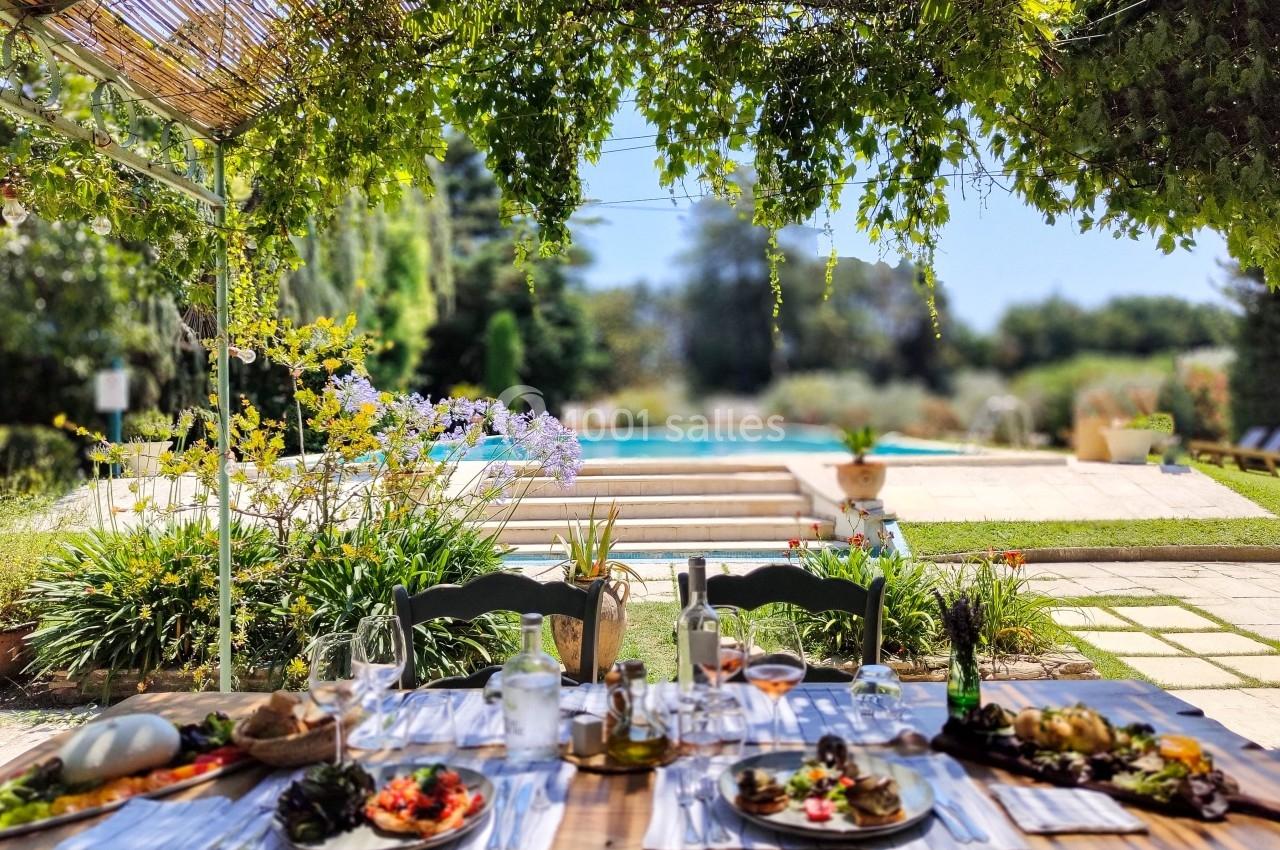 Table dressée pour un repas en plein air sous une pergola, avec vue sur un jardin verdoyant et une piscine.