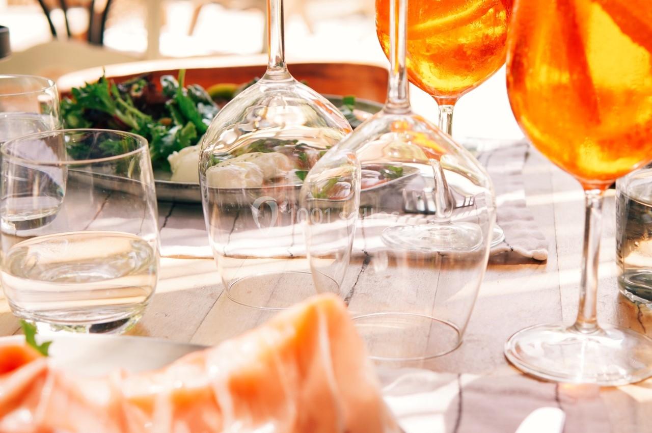 Table dressée en extérieur avec des verres de spritz, une assiette de jambon cru et une salade verte.
