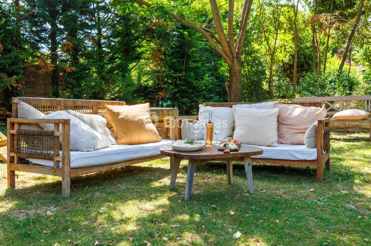 Canapé en bois avec coussins beiges et table basse sur une pelouse, entourés d'arbres dans un jardin ensoleillé.