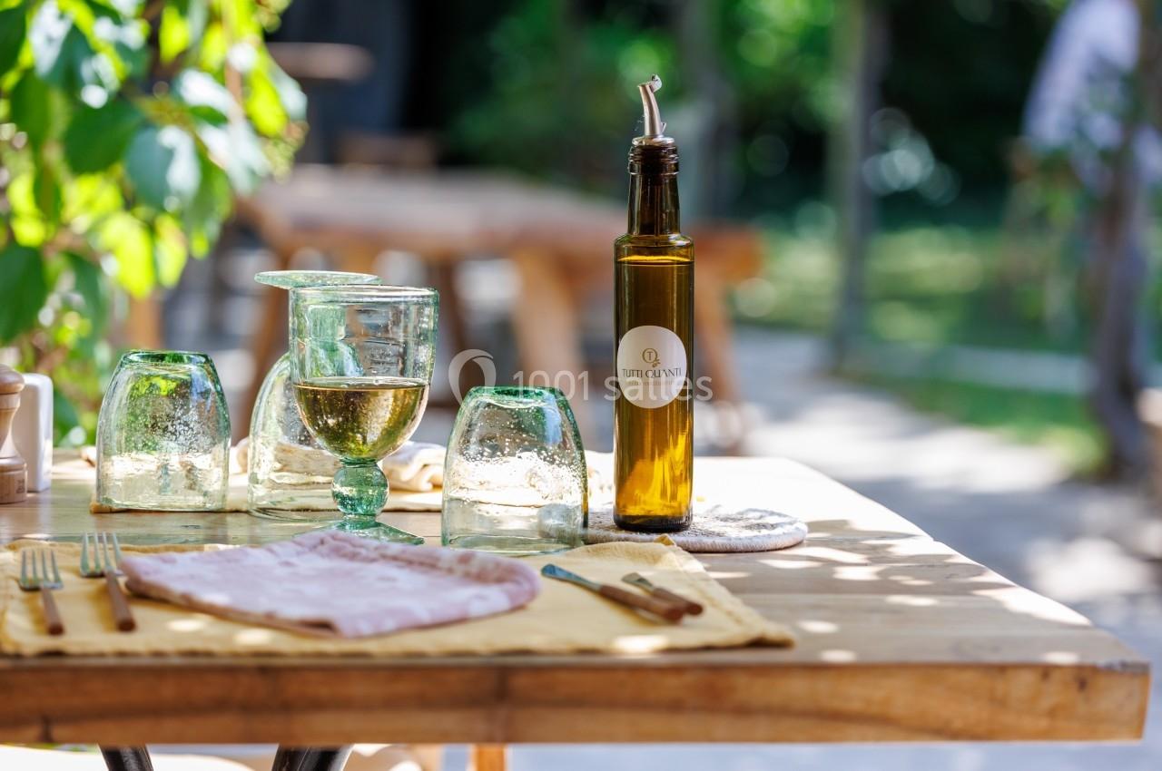Bouteille d'huile d'olive, verres et serviette disposés sur une table en bois dans un cadre extérieur verdoyant.
