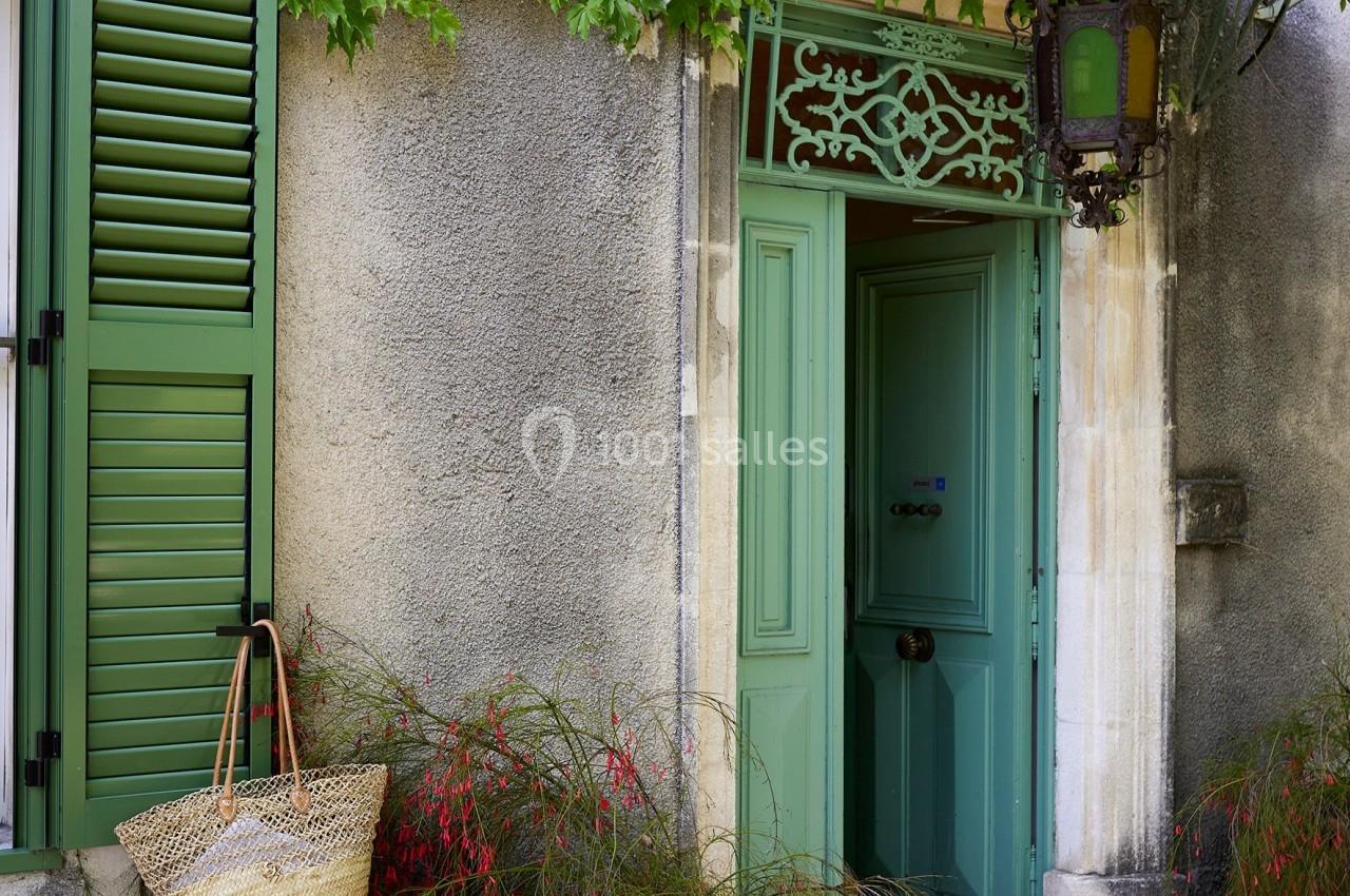 Entrée de maison avec porte et volets verts, plantes en pots et panier suspendu sous une marquise en métal.