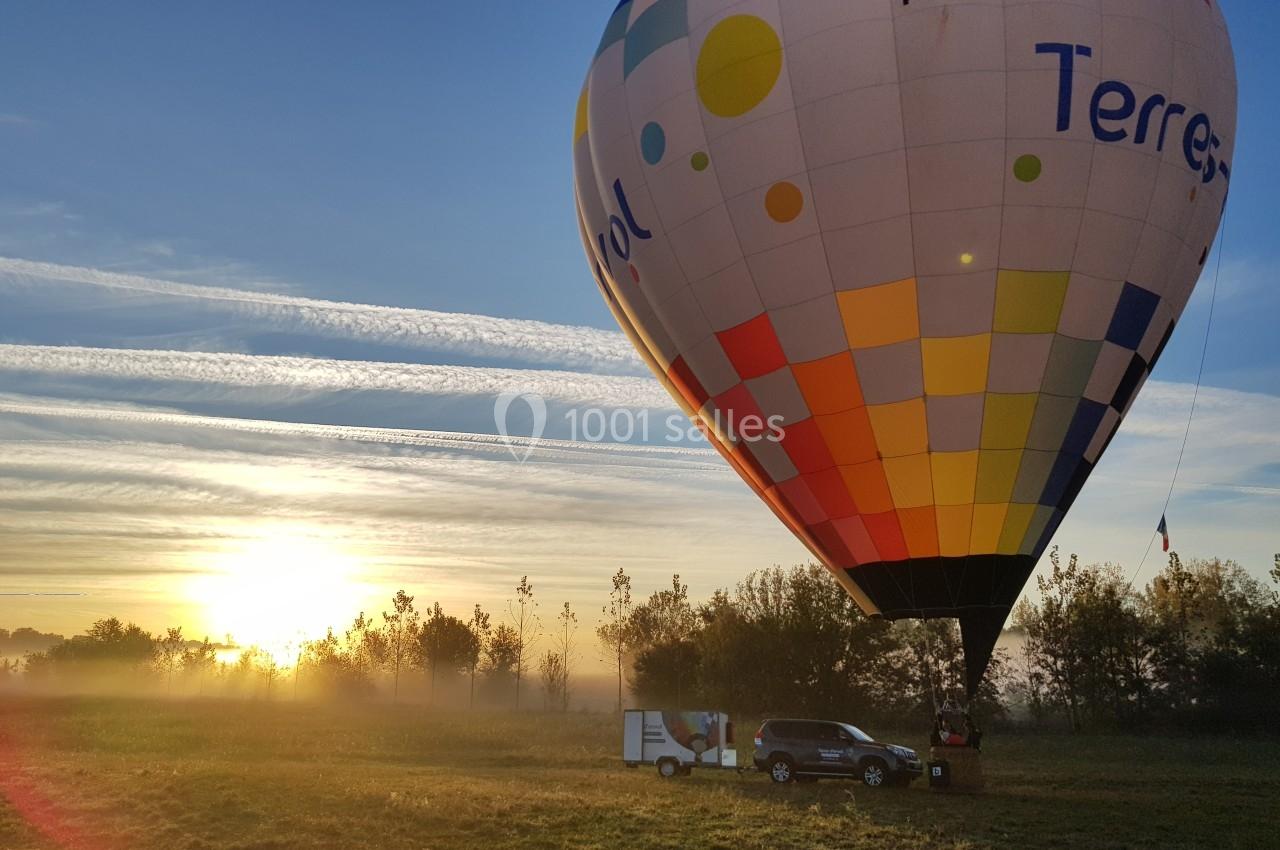 Montgolfière colorée en cours de gonflage dans un champ au lever du soleil, avec une voiture et une remorque à proximité.