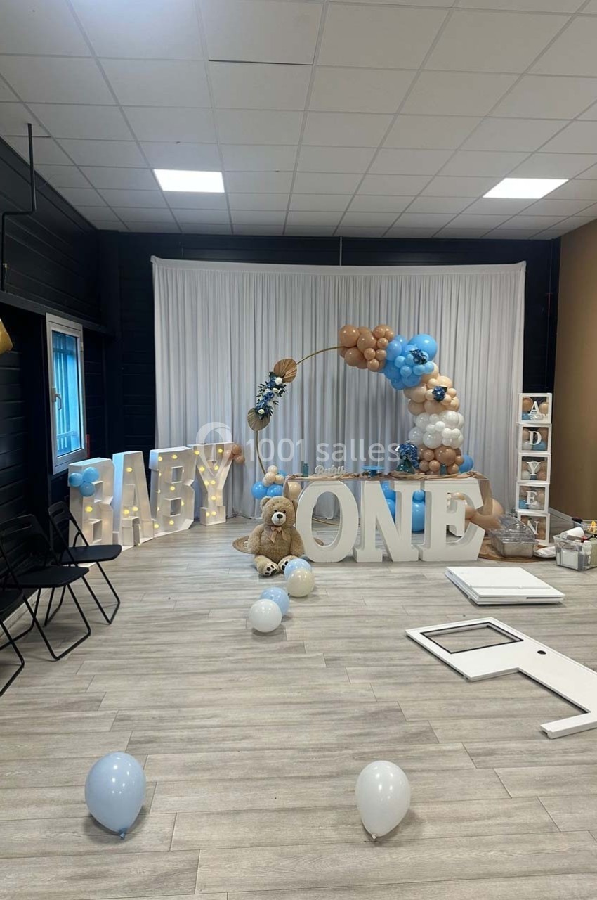Décoration de fête avec ballons bleus, blancs et beiges, lettres géantes ’BABY ONE’ et un ours en peluche.