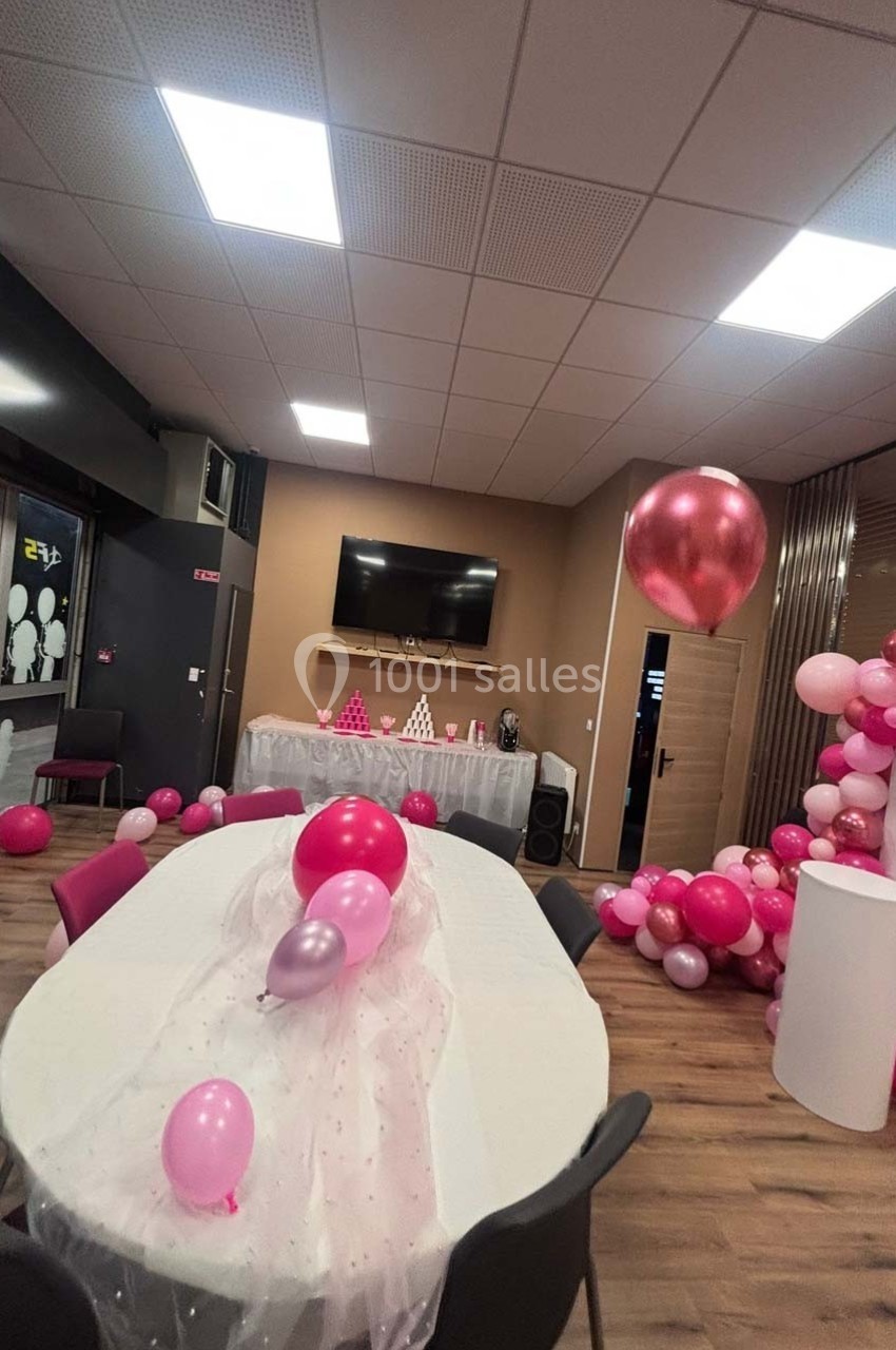 Salle décorée pour une fête, avec ballons roses et blancs, table ovale habillée de tulle et écran mural en arrière-plan.