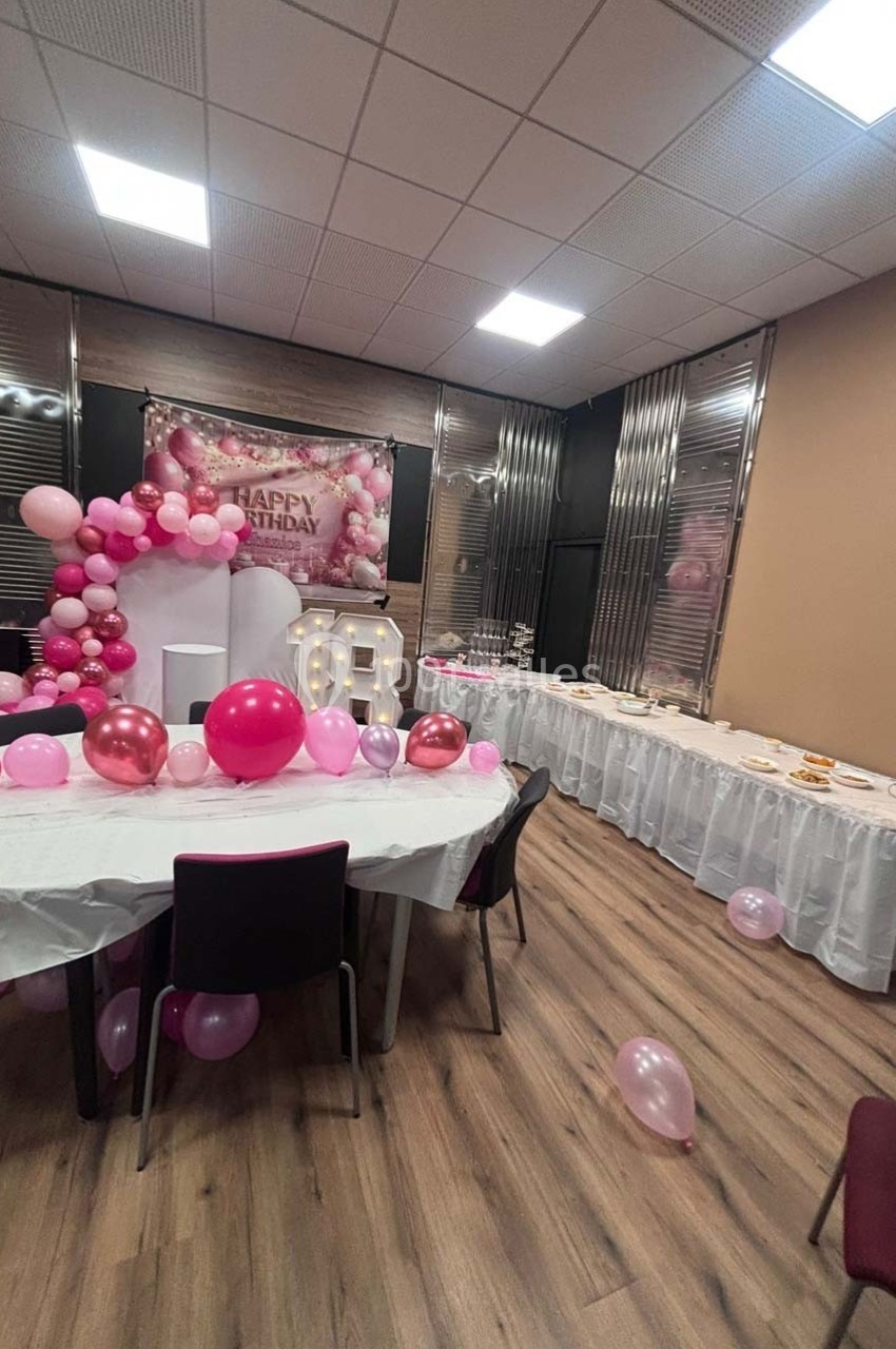 Salle décorée pour un anniversaire avec ballons roses, table dressée et panneau ’Happy Birthday’ en arrière-plan.