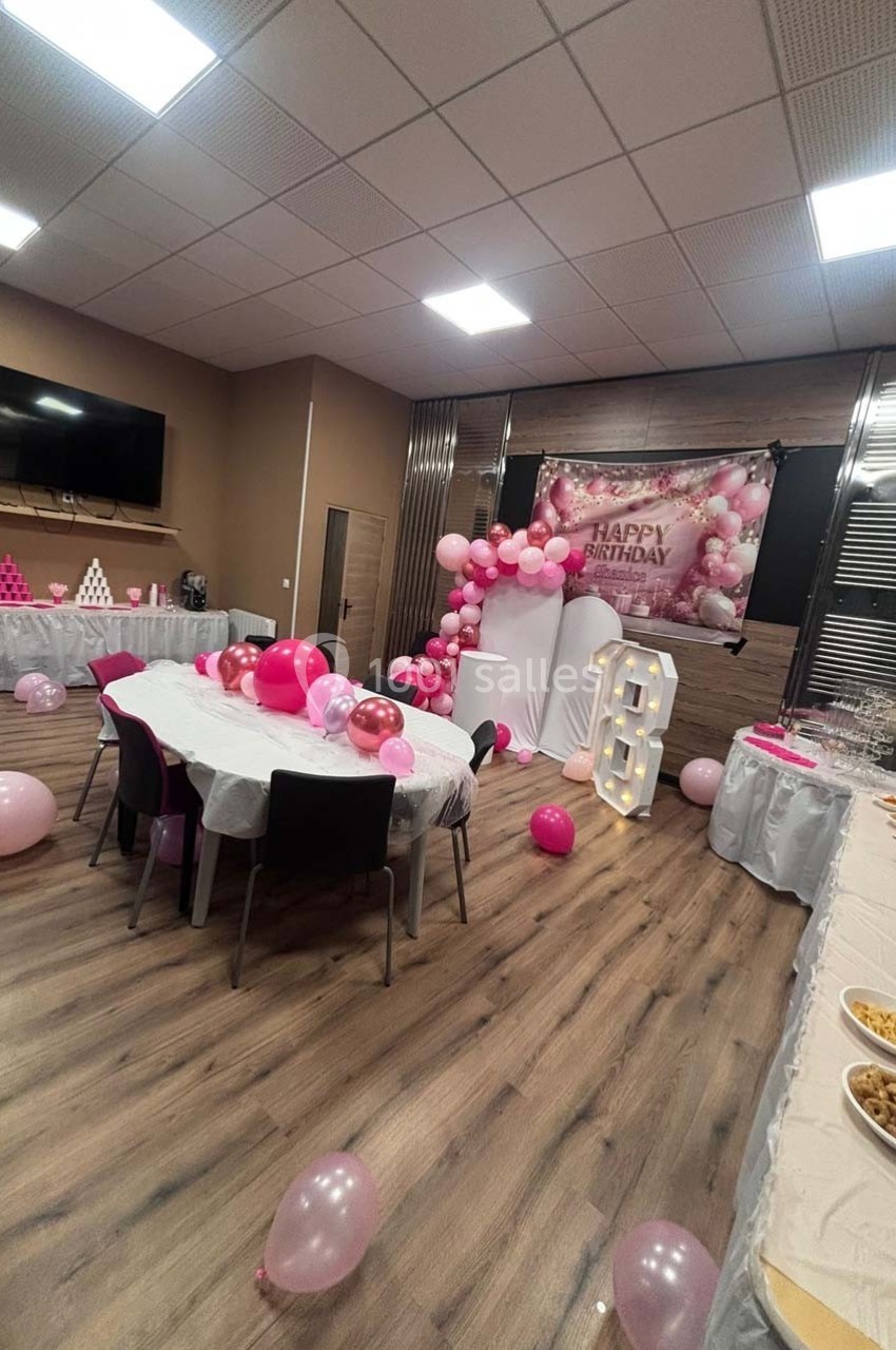 Salle décorée pour un anniversaire avec ballons roses, guirlandes, tables dressées et un panneau ’Happy Birthday’.