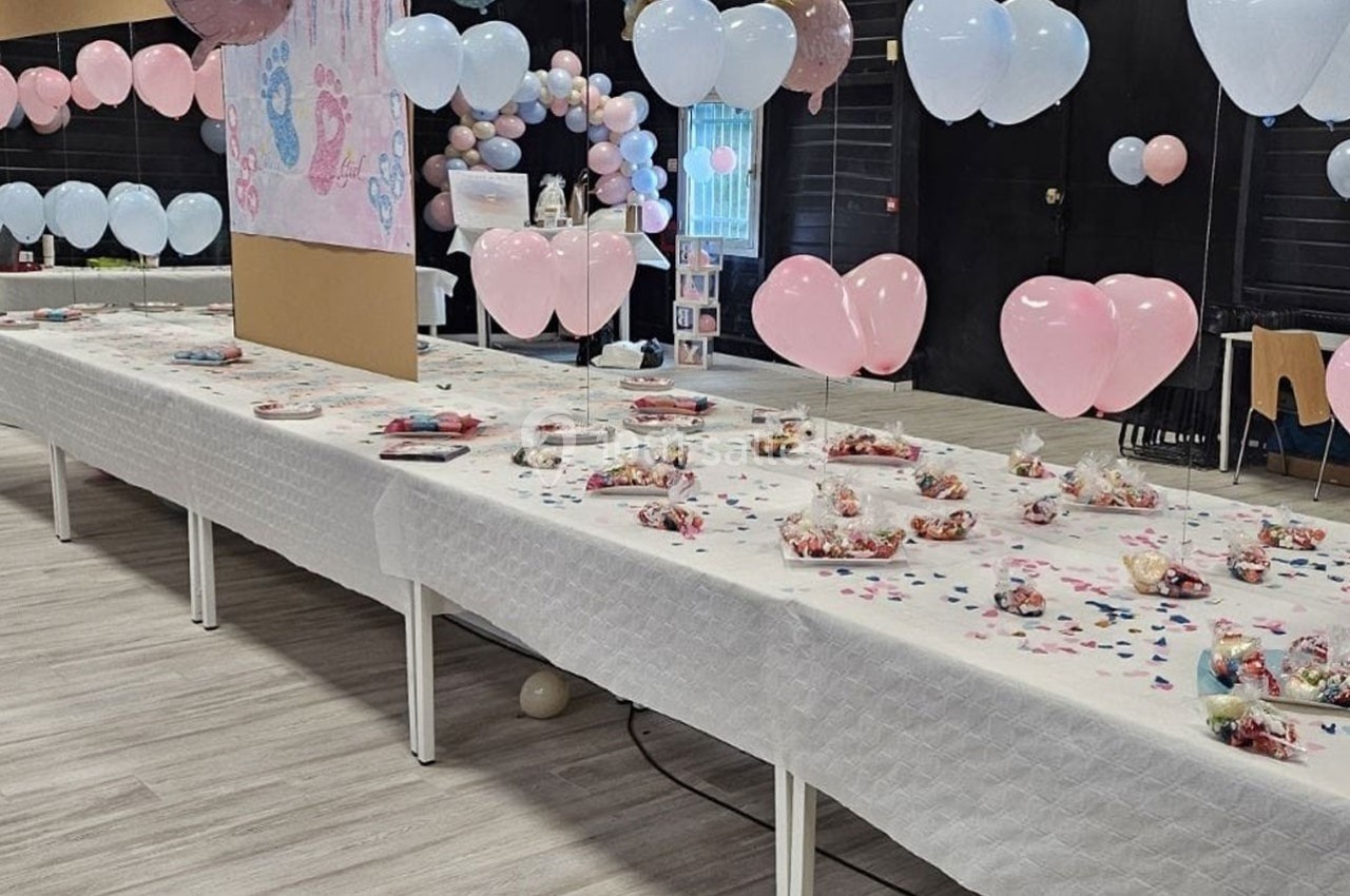 Salle décorée pour une fête avec ballons roses et bleus, tables dressées avec confiseries et confettis.