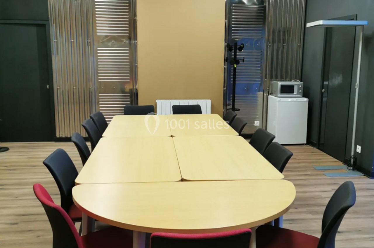 Salle de réunion avec une grande table en bois entourée de chaises noires et rouges, sol en parquet et murs métalliques.
