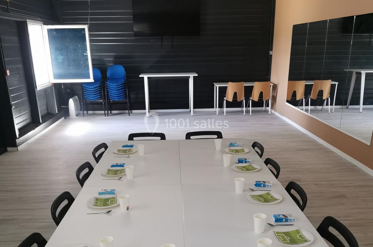 Salle avec une grande table dressée pour un repas, chaises empilées, tableau blanc et écran mural en arrière-plan.