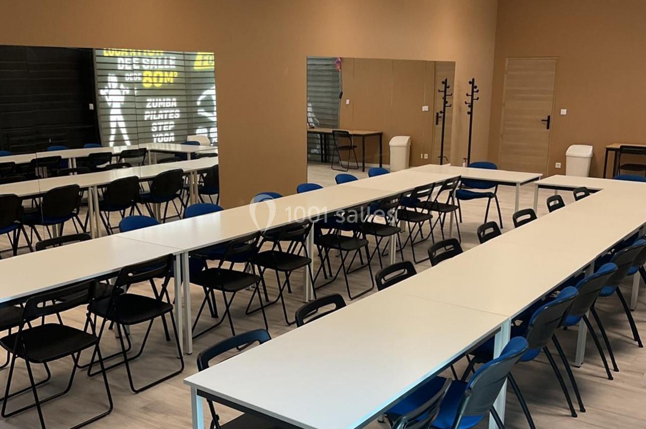 Salle de réunion avec tables blanches alignées en U, chaises noires et bleues, murs beige et miroir au fond.