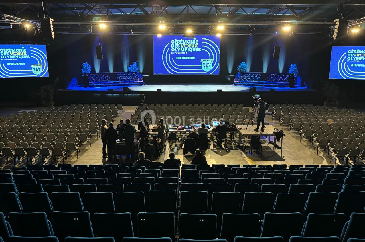 Salle de spectacle avec scène éclairée, écrans affichant ’Cérémonie des Vœux Olympiques’, et rangées de sièges vides.