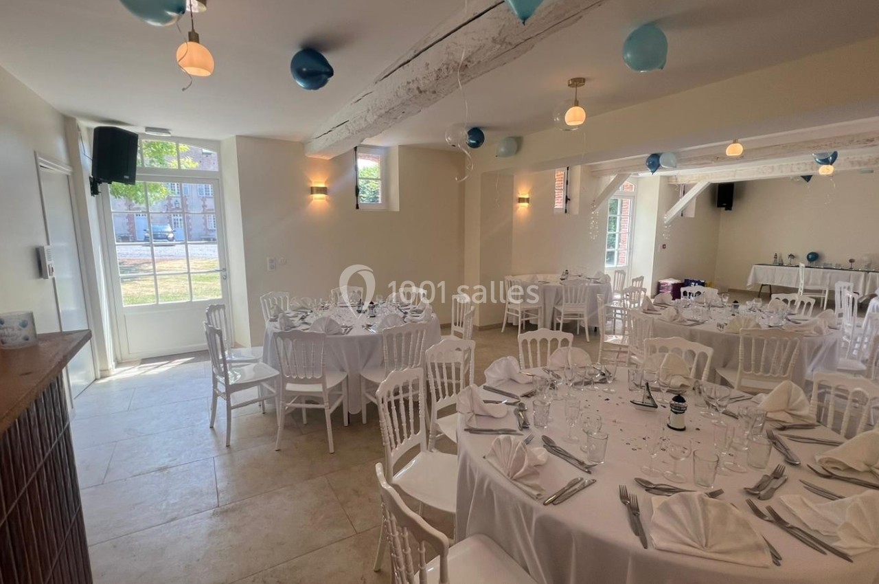 Salle de réception lumineuse avec tables rondes dressées, chaises blanches et décorations suspendues.