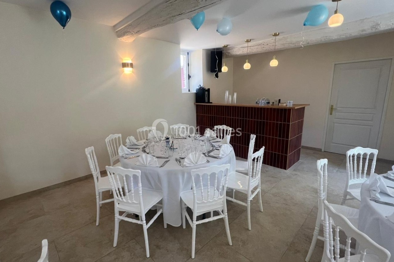 Salle décorée avec des ballons bleus et blancs, tables rondes dressées avec nappes blanches et chaises assorties.