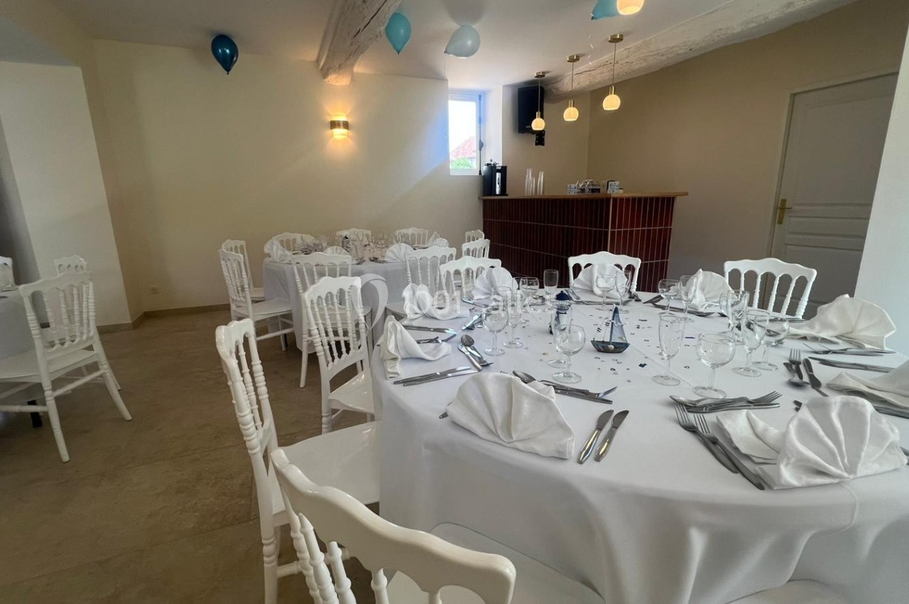 Salle de réception lumineuse avec tables rondes dressées, nappes blanches, serviettes pliées et chaises assorties.