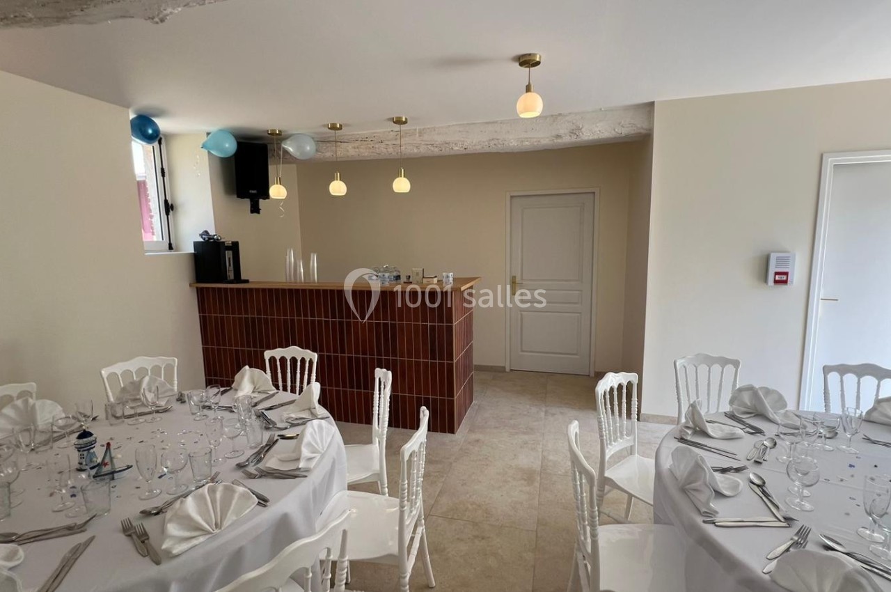 Salle de réception lumineuse avec tables dressées, chaises blanches et un bar en bois en arrière-plan.