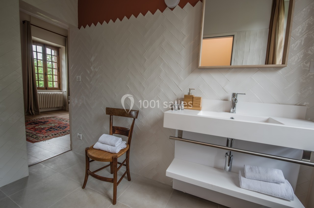 Salle de bain lumineuse avec lavabo moderne, chaise en bois et vue sur une pièce adjacente avec fenêtre.