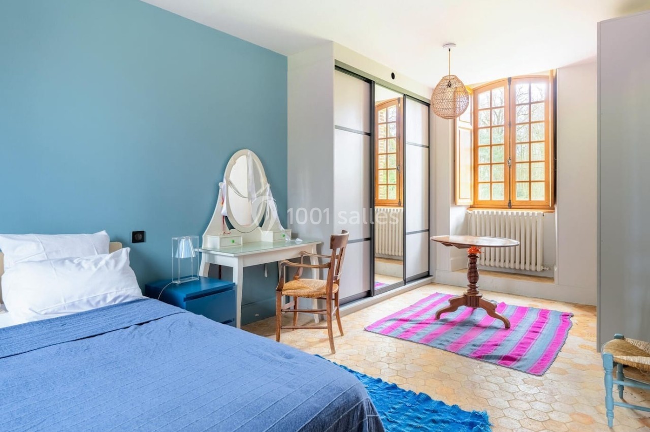 Chambre lumineuse avec lit, coiffeuse, table ronde, tapis coloré et grande fenêtre laissant entrer la lumière naturelle.
