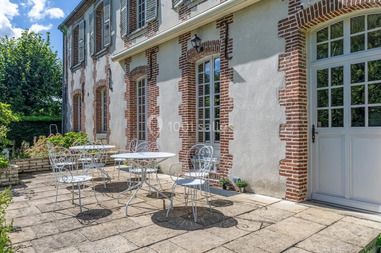 Terrasse en pierre avec tables et chaises en fer forgé, adossée à une maison en briques et crépi, entourée de verdure.
