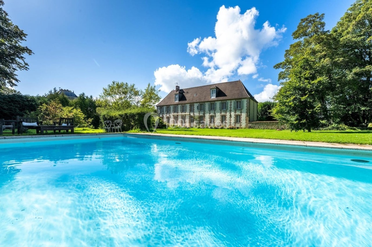 Piscine extérieure entourée de verdure avec une maison en pierre et un ciel bleu en arrière-plan.