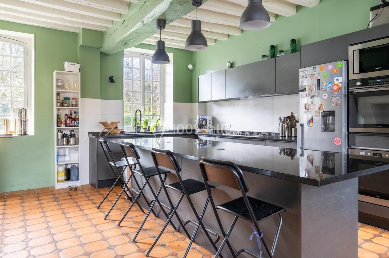 Cuisine lumineuse avec murs verts, îlot central noir, tabourets pliants et électroménagers intégrés.