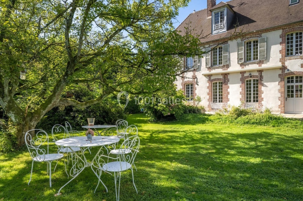 Table et chaises blanches en fer forgé disposées sous un arbre dans le jardin d'une maison en briques et pierres.