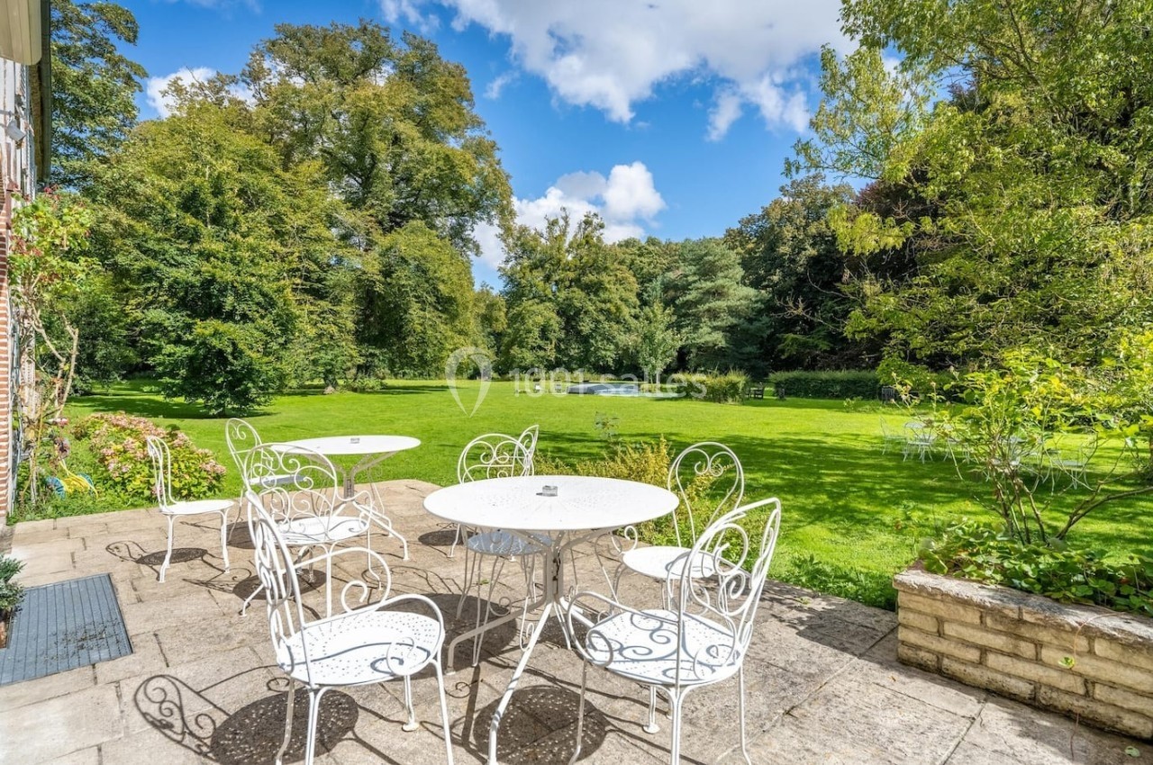 Terrasse avec tables et chaises blanches en fer forgé, donnant sur un grand jardin verdoyant parsemé d'arbres.