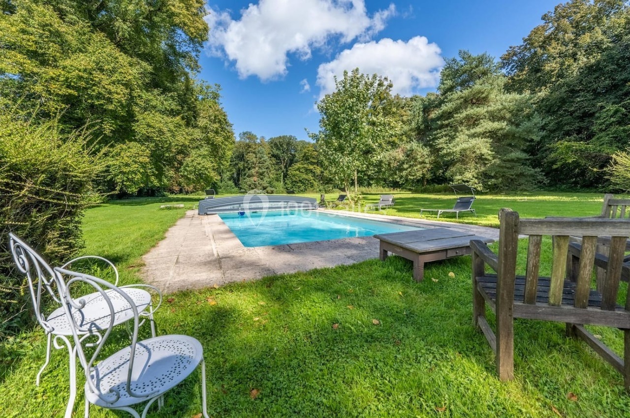 Piscine extérieure entourée de chaises, bancs et végétation luxuriante sous un ciel bleu.