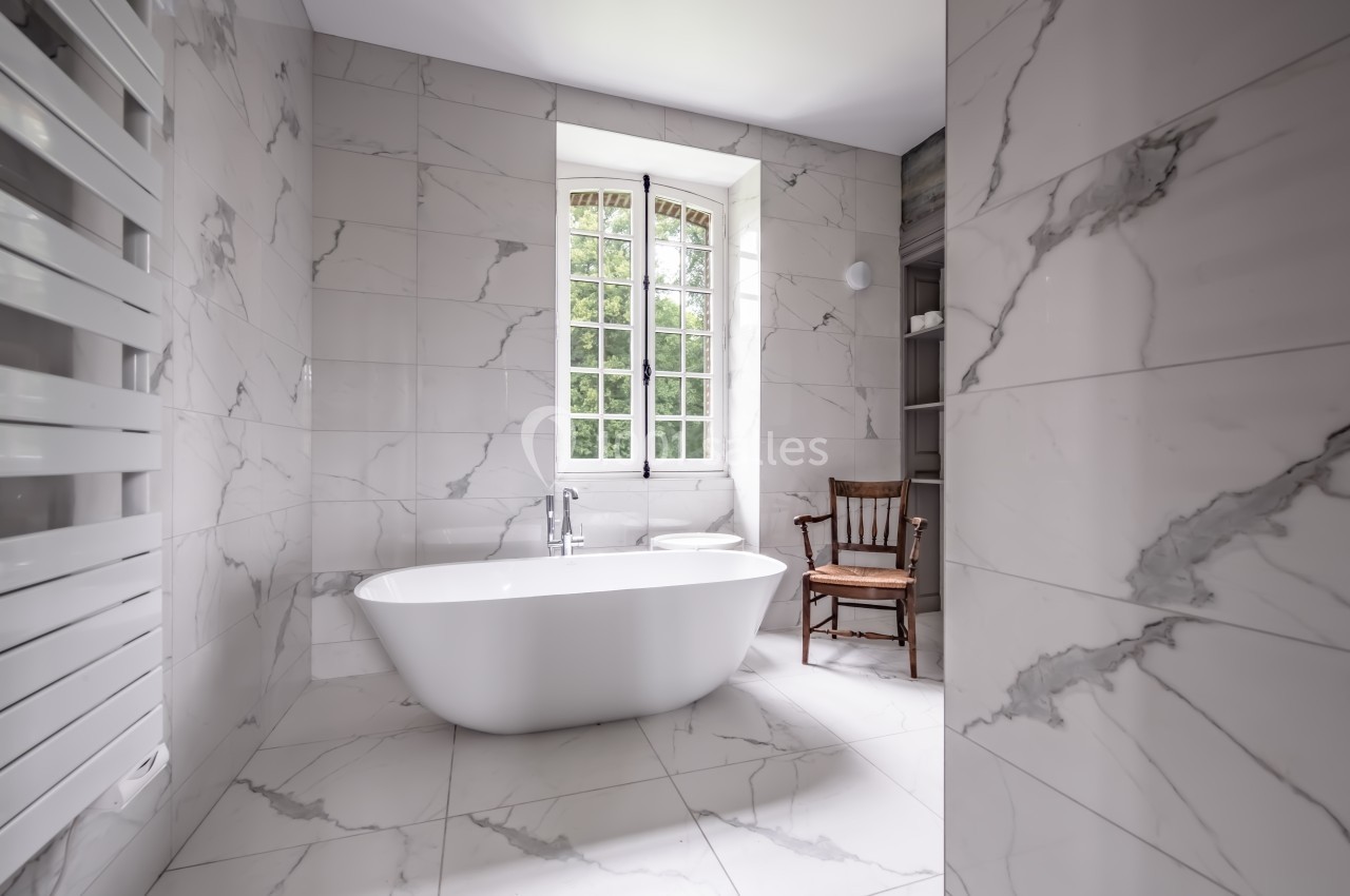 Salle de bain lumineuse avec baignoire îlot, murs en carrelage effet marbre, fenêtre et chaise en bois.