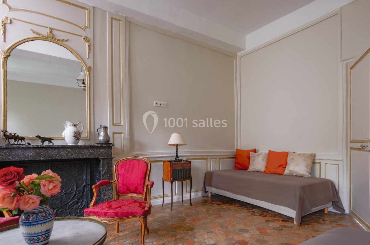 Salon élégant avec cheminée en marbre, fauteuil rouge, table basse avec fleurs et canapé beige orné de coussins colorés.